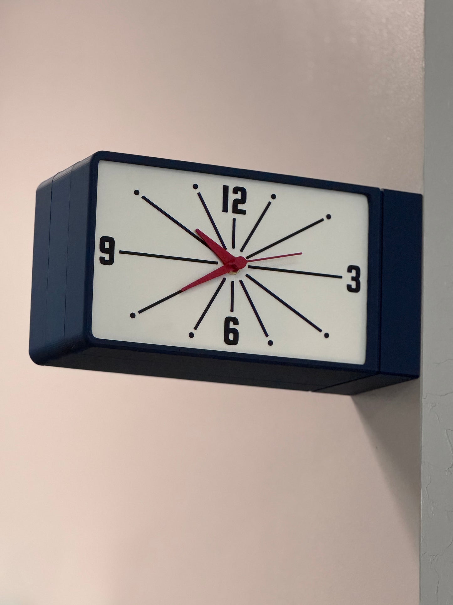 Rectangular wall clock with dark blue frame and red hands on a plain wall.